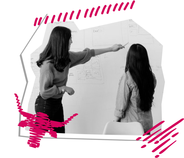 The teacher, wearing a blouse and jeans, is pointing to the white board. The student, in a jean jacket and with long hair, is looking on.