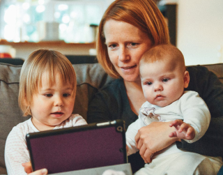 A person holding a baby and sitting with a toddler. All three are looking at a tablet and exploring the content.