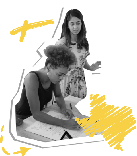 Two students are working on a project. One student stands next to a desk. The other student is leaning over a large sheet of paper on the desk and writing