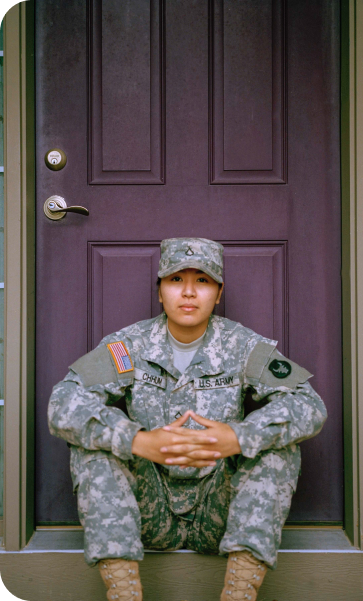 A person in Army fatigues sitting in front of a purple door. Their hands are clasped in front of them, and they look straight into the camera.